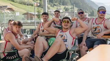 Eine Gruppe junger Männer sitzt entspannt an einem Tisch im Freien. Sie tragen sportliche Shirts und Sonnenbrillen und scheinen Spaß zu haben.