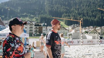 Zwei Männer stehen am Strand und schauen in die Ferne. Im Hintergrund sind Berge und Gebäude zu sehen.