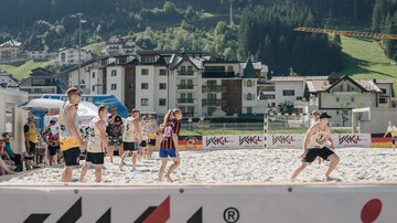 Gruppenspiele am Strand mit mehreren Kindern. Im Hintergrund sind Berge und moderne Gebäude zu sehen.