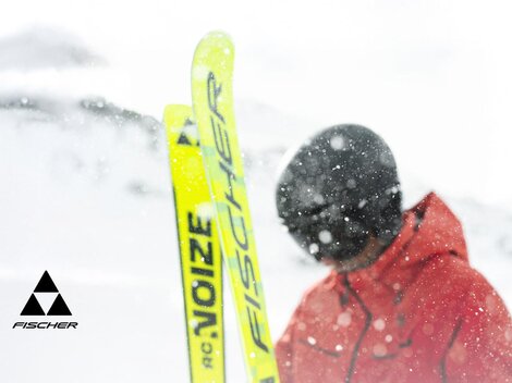 Ein Skifahrer in einem roten Anzug steht im Schnee. Die Umgebung ist winterlich und verschneit, während die Skier leuchtend gelb sind.