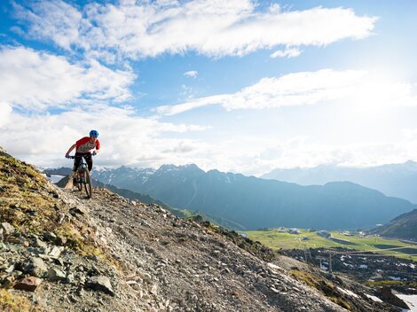 Ein Mountainbiker fährt über einen steilen, steinigen Pfad mit spektakulärer Berglandschaft im Hintergrund. Der Himmel ist klar und es gibt viele Wolken.