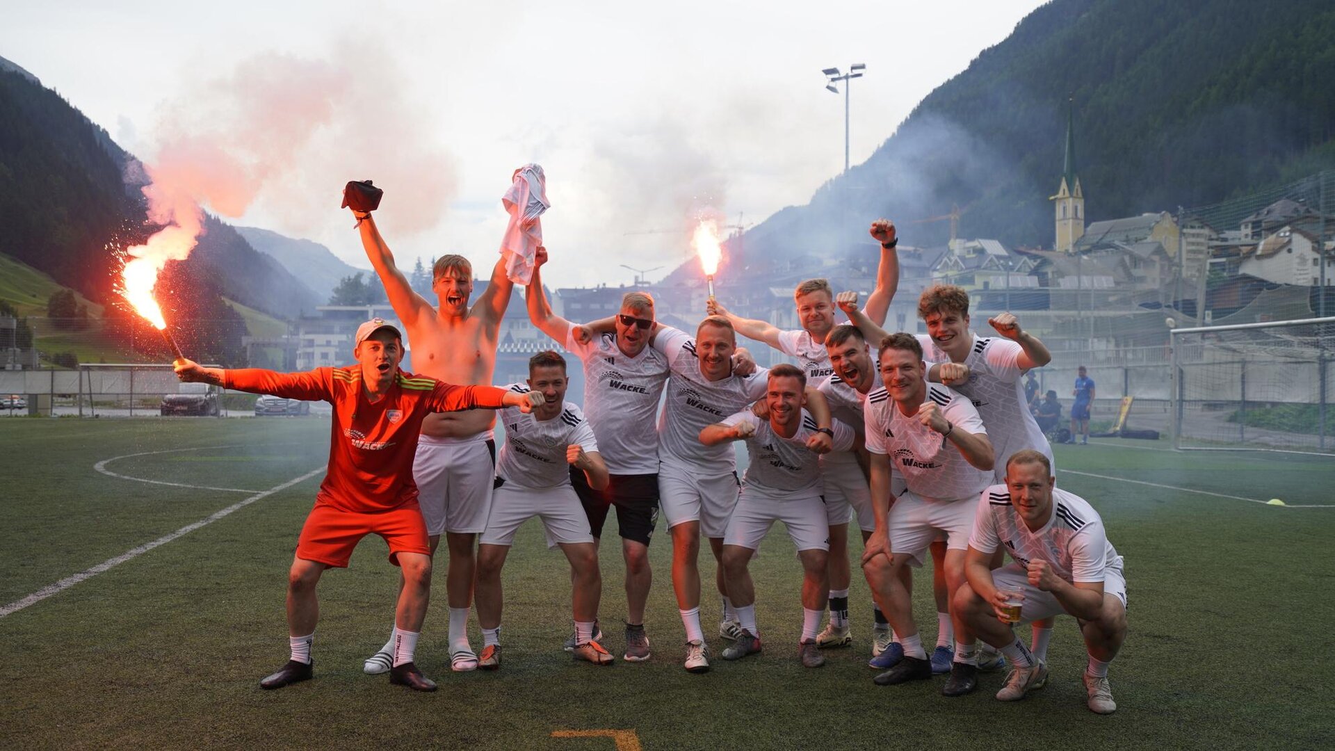 Eine Gruppe von Fußballspielern feiert auf dem Platz. Sie sind fröhlich und halten Fackeln in der Hand, während im Hintergrund eine malerische Berglandschaft zu sehen ist.