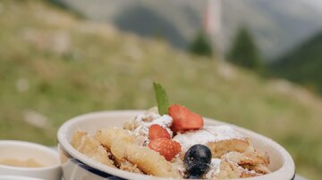 Ein köstliches Gericht mit gebratenen Apfelstücken, frischen Früchten und Puderzucker. Dazu gibt es zwei kleine Schalen mit Marmelade und Sahne.