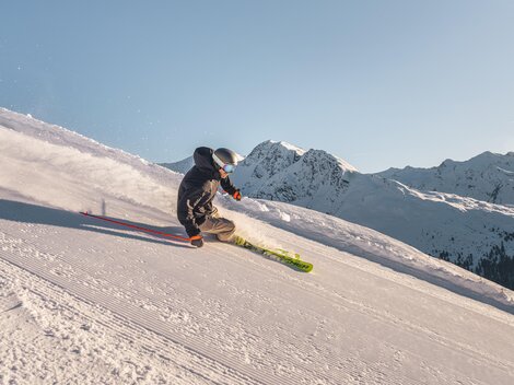Ein Skifahrer fährt eine verschneite Piste hinunter. im Hintergrund sind schneebedeckte Berge zu sehen.