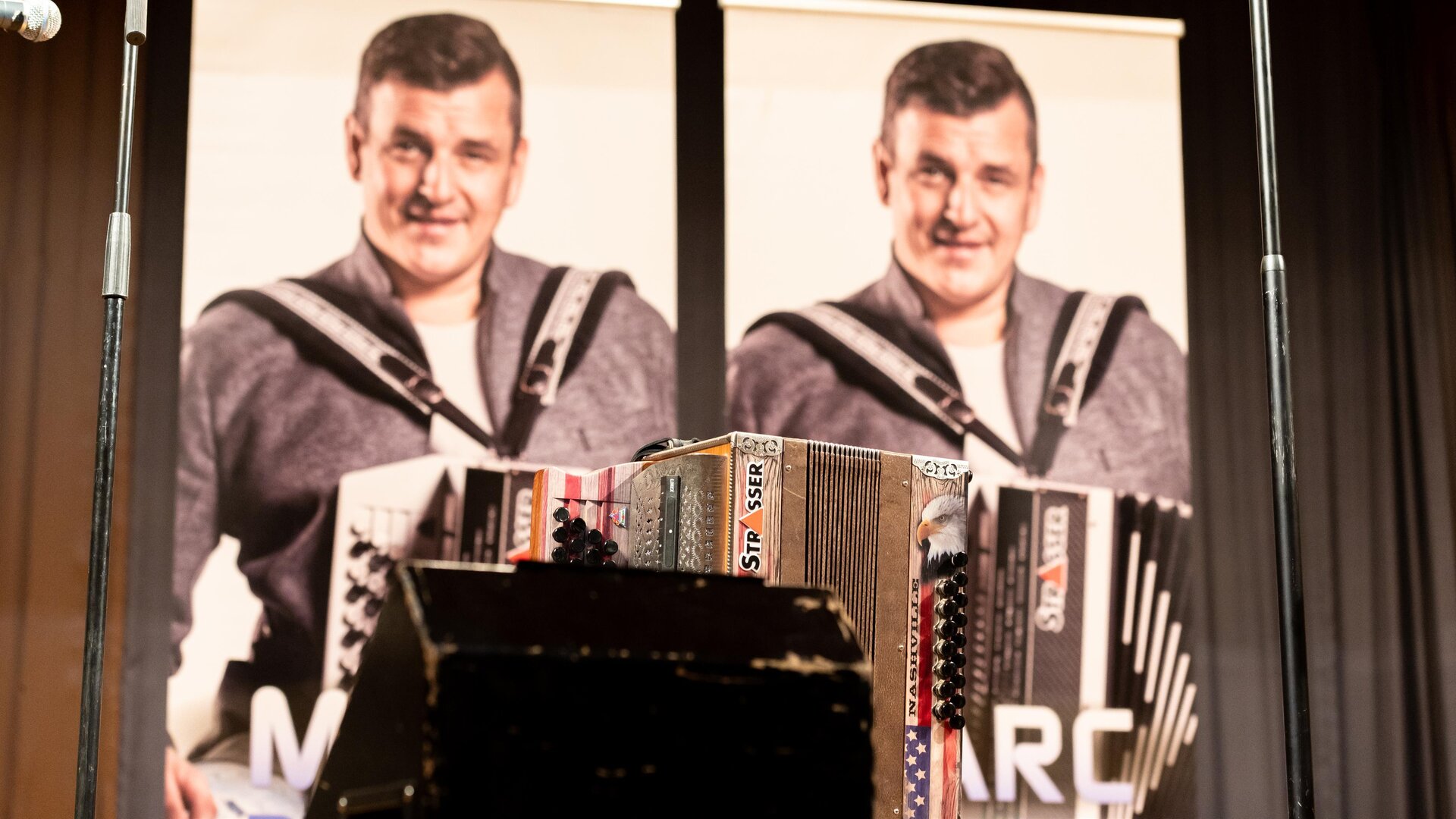 Zwei Plakate mit einem lächelnden Mann, der ein Akkordeon hält. Im Vordergrund ist ein weiteres Akkordeon sichtbar.