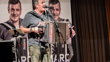 Ein Musiker spielt Akkordeon auf einer Bühne. Im Hintergrund sind Plakate mit seinem Bild zu sehen.