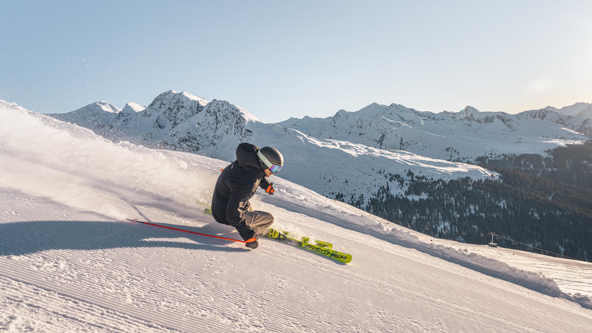 Ein Skifahrer fährt über eine schneebedeckte Piste in den Bergen. Im Hintergrund sind majestätische Berge und ein klarer Himmel zu sehen.