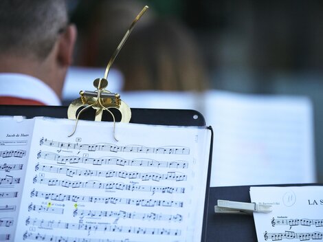 Ein Ausschnitt von Notenblättern auf einem Notenständer. Im Hintergrund sind Musiker zu sehen, die sich auf eine Aufführung vorbereiten.