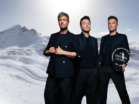Drei Männer stehen vor einer verschneiten Berglandschaft. Sie tragen elegante schwarze Kleidung und blicken in die Kamera.