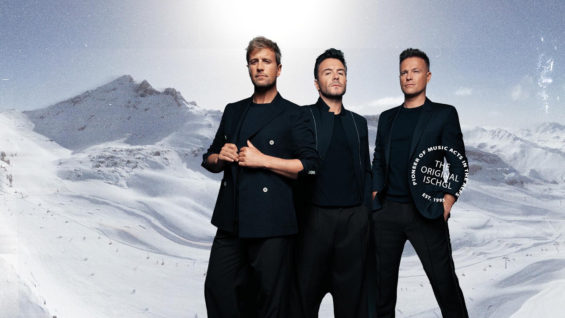 Drei Männer stehen vor einer verschneiten Berglandschaft. Sie tragen elegante schwarze Kleidung und blicken in die Kamera.