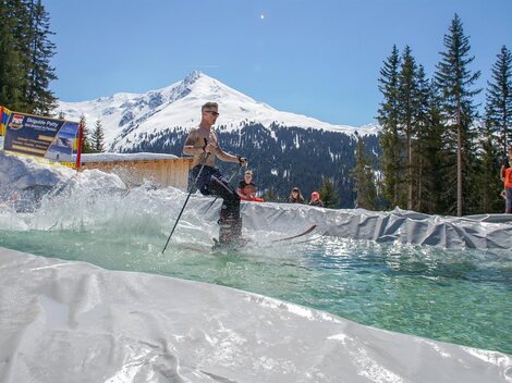 Ein Skifahrer fährt über einen Wassersprung in einer alpinen Umgebung. Im Hintergrund sind schneebedeckte Berge und Tannenbäume zu sehen.