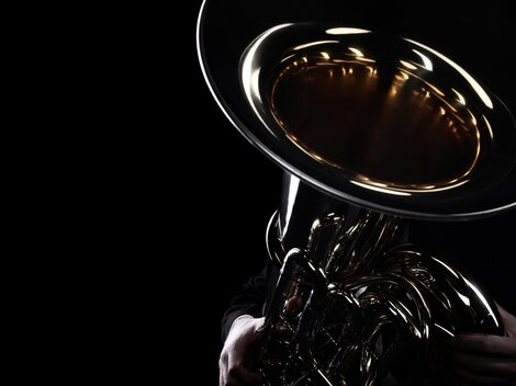 Ein Musiker hält eine glänzende Tuba im Dunkeln. Nur der untere Teil des Instruments und die Hände sind sichtbar.