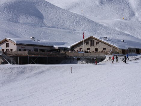Eine Berghütte im Schnee mit skifahrenden Personen. Die Landschaft ist von schneebedeckten Bergen umgeben.