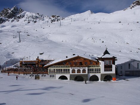 Eine schneebedeckte Landschaft mit Bergmassiven im Hintergrund. Im Vordergrund steht ein traditionelles Berghaus.