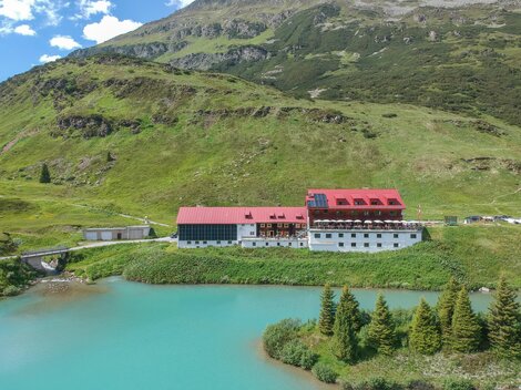 Ein malerisches Hotel am Ufer eines klaren, blauen Sees, umgeben von grünen Hügeln. Im Hintergrund sind Berge und ein blauer Himmel zu sehen.