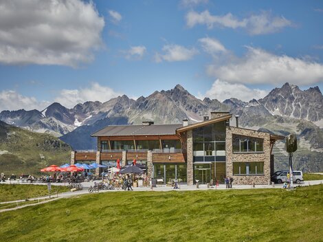 Ein modernes Gebäude in einer malerischen Berglandschaft. Die Terrasse ist mit Gästen und bunten Sonnenschirmen geschmückt.