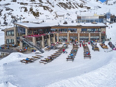 Ein modernes Berghaus umgeben von schneebedeckten Bergen. Viele Liegen stehen bereit, um die Sonne zu genießen.