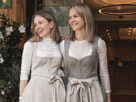 Zwei Frauen stehen lächelnd vor einem Gebäude. Beide tragen traditionelle Kleider mit modernen Akzenten.