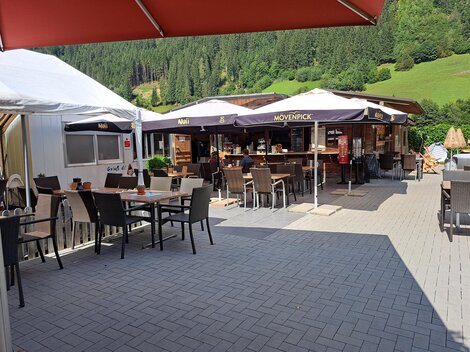 Ein gemütlicher Außenbereich eines Restaurants mit Tischen und Stühlen. Im Hintergrund befindet sich das Restaurant mit Schirmen und einer grünen Landschaft.