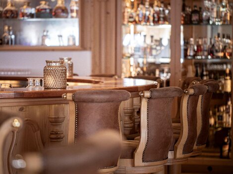 Eine stilvolle Bar mit holzverkleideten Stühlen und eleganten Gläsern. Im Hintergrund sind Regale mit verschiedenen Spirituosen sichtbar.
