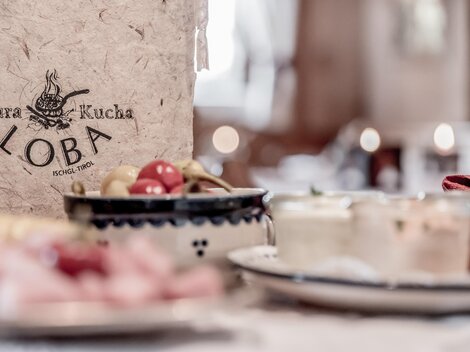 Ein dekoratives Restaurantmenü mit dem Namen "Loba" ist im Vordergrund zu sehen. Im Hintergrund befinden sich verschiedene kleine Gerichte und Snacks.