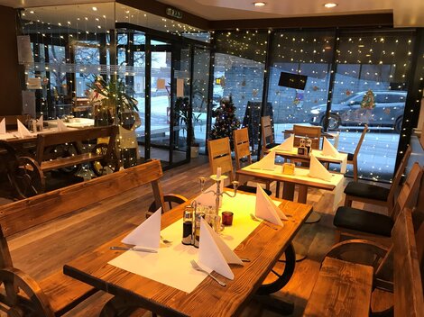 Ein gemütliches Restaurant mit Holztischen und -stühlen. Durch große Fenster sieht man weihnachtliche Dekorationen und stimmungsvolles Licht.