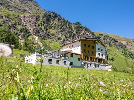 Ein großes Gebäude in den Bergen, umgeben von grünen Wiesen und Felsen. Der Himmel ist klar und blau.