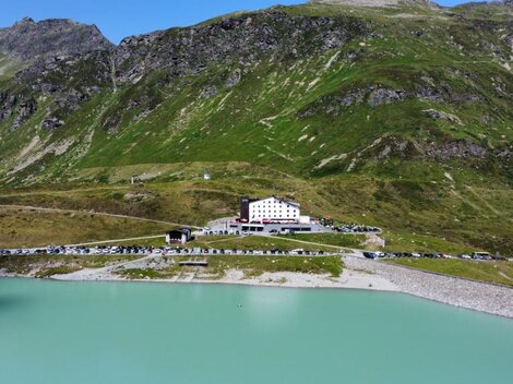 Ein malerischer Bergsee mit hellem, türkisfarbenem Wasser. Am Ufer steht ein großes, weißes Gebäude und grünes Gebirge umgibt die Landschaft.