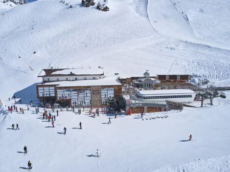 Ein Skigebiet mit viel Schnee und einem großen Holzgebäude. Zahlreiche Skifahrer und Snowboarder genießen die winterliche Landschaft.