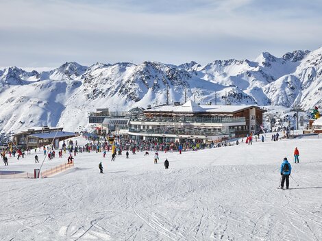 Ein belebtes Skigebiet mit vielen Menschen auf der Piste. Im Hintergrund sind schneebedeckte Berge und ein modernes Gebäude zu sehen.