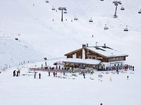 Ein Holzhaus in den Bergen, umgeben von Schnee und Skifahrern. Im Hintergrund sind Skilifte zu sehen.