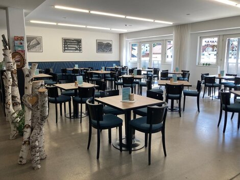 Ein modernes Restaurant mit Tischen und Stühlen. Die Wände sind hell und große Fenster lassen viel Licht herein.