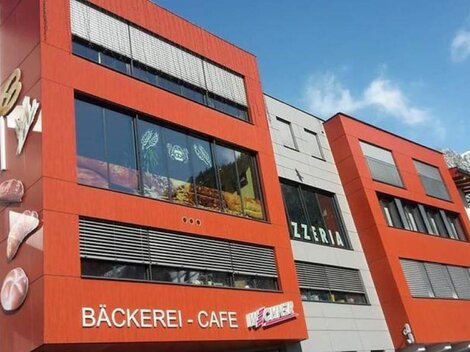 Ein modernes Gebäude mit roter Fassade und großen Fenstern. Darin sind ein Bäckerei-Café und eine Pizzeria angesiedelt.