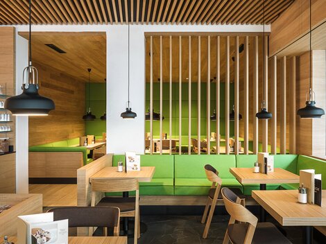 Ein modernes Café mit Holzmöbeln und einem klaren, minimalistischen Design. Die Wände sind mit einem lebendigen Grün akzentuiert, und es gibt stilvolle Pendelleuchten.