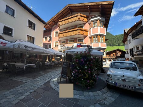 Eine malerische Stadtstraße mit charmanten Gebäuden und einem kleinen Platz. Auf dem Platz stehen Blumen und ein klassisches Auto parkt in der Nähe.