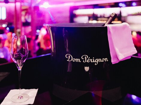Eine Flasche Dom Pérignon in einem eleganten Eiskübel steht bereit. Im Hintergrund ist eine festliche Atmosphäre mit einem unscharfen Publikum zu sehen.
