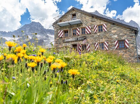 Eine malerische Berghütte aus Stein mit bunten Fensterläden. Der Garten ist mit gelben Blumen und einer beeindruckenden Bergkulisse umgeben.