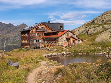 Eine malerische Berghütte inmitten einer grünen Landschaft. Im Vordergrund befindet sich ein kleiner Teich und im Hintergrund sind beeindruckende Berge zu sehen.