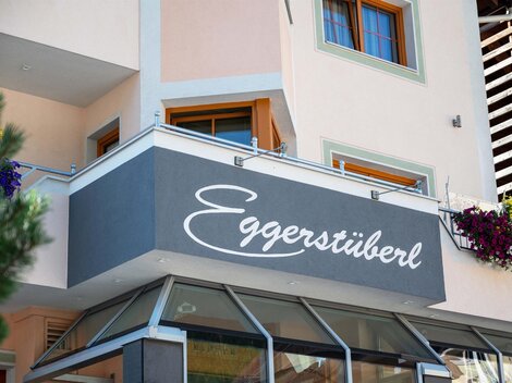 Ein modernes Gebäude mit einer Strickfront und einem großen Schild mit der Aufschrift „Eggerstüberl“. Blumen schmücken die Fenster und verleihen dem Ort eine einladende Atmosphäre.