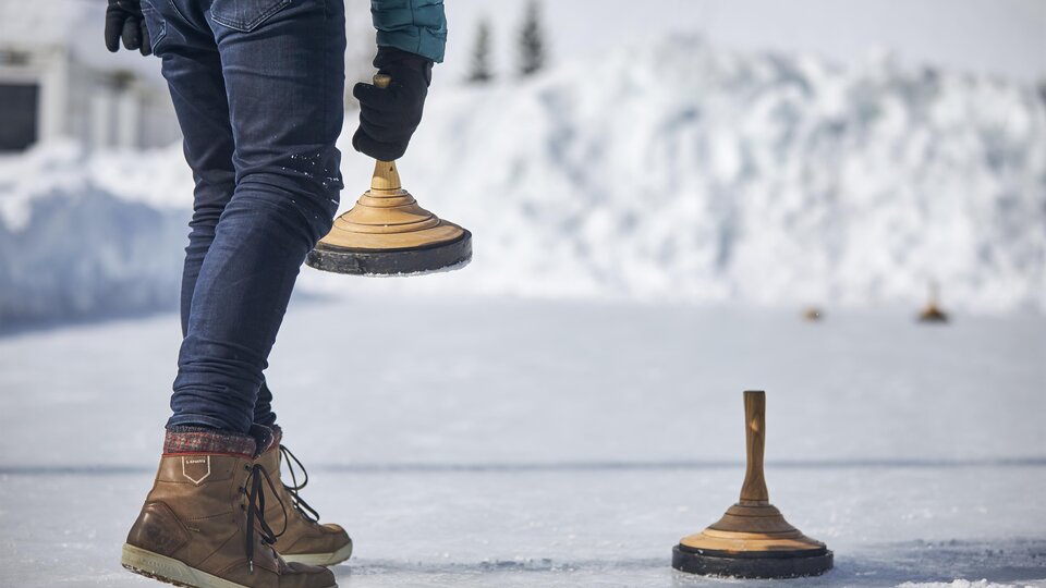 Eine Person steht auf einer gefrorenen Fläche und hält einen Holzblock in der Hand. Im Hintergrund sind schneebedeckte Hügel zu sehen.