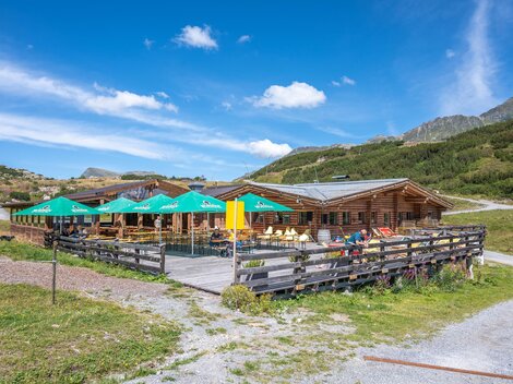 Eine gemütliche Berghütte mit Terrasse und Sonnenschirmen. Umgeben von grünen Wiesen und Bergen unter einem blauen Himmel.