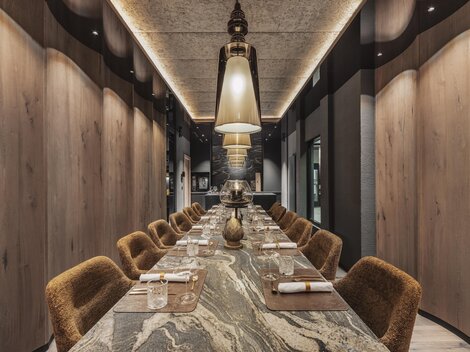 Ein eleganter Restaurantbereich mit einem langen Tisch und gemütlichen Stühlen. Die Atmosphäre ist modern und stilvoll, beleuchtet von großen Lampen.