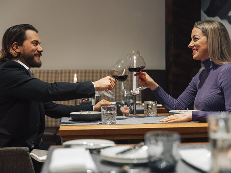 Ein elegantes Dinner-Paar stoßen mit Rotwein an. Der Tisch ist gedeckt und die Atmosphäre wirkt gemütlich.