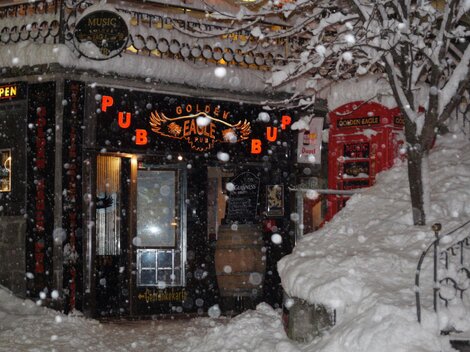 Ein gemütlicher Pub mit dem Namen "Golden Eagle" ist im Schnee verborgen. Die Schneeflocken fallen dicht, während das warme Licht des Eingangs einladend scheint.