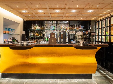 Eine elegante Bar mit einer goldenen Theke und einer Vielzahl von Spirituosen. Die Wandregale sind mit Gläsern und Getränken dekoriert.