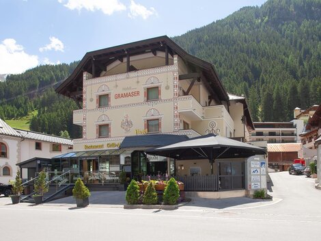 Ein malerisches Hotel im gebirgigen Umfeld mit grünen Bergen im Hintergrund. Vor dem Hotel gibt es eine gepflasterte Fläche mit Tischen und Stühlen.