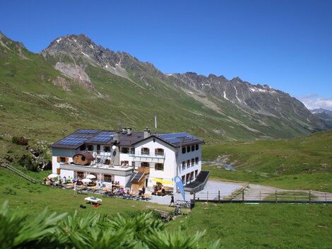 Ein charmantes Berghaus in einer grünen Landschaft. Im Hintergrund sind majestätische Berge und blauer Himmel zu sehen.