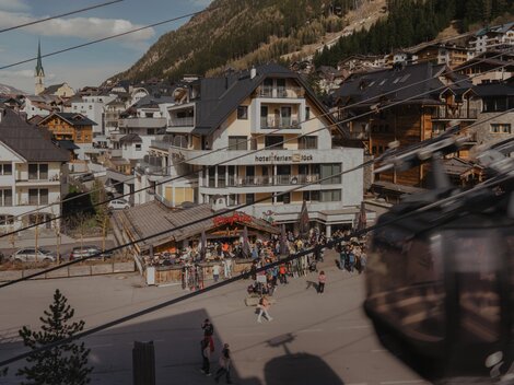 Ein belebter Ort in den Bergen mit einem Hotel und vielen Menschen. Im Hintergrund sind Holzgebäude und eine Seilbahn zu sehen.