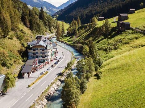 Eine malerische Landschaft mit einem Fluss, der durch ein Tal fließt. Am Ufer stehen ein paar Häuser und die Umgebung ist von grünen Wiesen und bewaldeten Hügeln umgeben.