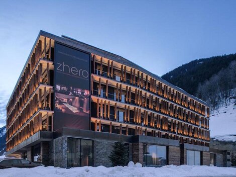 Ein modernes Hotel mit einer stilvollen Fassade und beleuchteten Balkonen. Umgeben von schneebedeckten Bergen und einem klaren Abendhimmel.